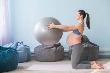 Pregnant european woman performs exercises with fitness ball. Expectant mother is doing yoga in the...