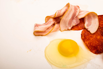 A broken chicken egg without a shell, slices of sausage and bacon and tomatoes on a white background. Close up.