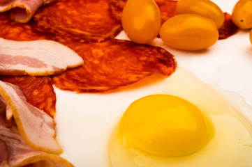A broken chicken egg without a shell, slices of sausage and bacon and tomatoes on a white background. Close up.