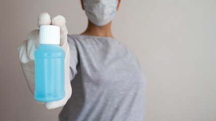 A woman with a surgical mask holds a plastic bottle of alcohol gel in her hand that is in a rubber glove.