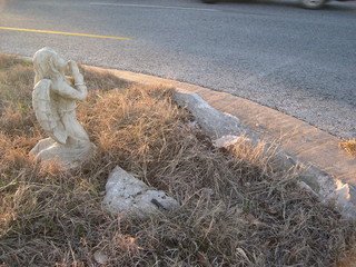Roadside traffic fatality memorial with broken curb and stone angel at sunset