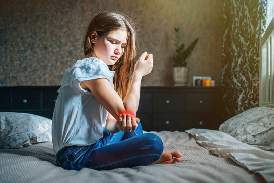 young brunette girl sits on a bed in her room, holds a sore elbow with her hand