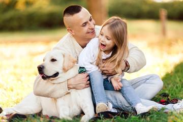 Happy father and daughter with dog labrador is having fun are sitting on green grass in park.