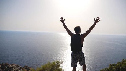 Male hiker standing on peak and victoriously raising hands rejoicing achievement or success. Young man actively resting during trip enjoying freedom at hill top. Scenic background. Dolly shot Slow mo