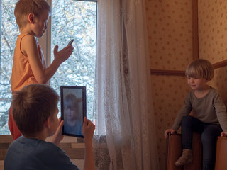 Three boys are playing with tablet and smartphone on leather sofa in bedroom by window. brothers spend time together in quarantine in rural house. Older children take pictures of young and show photos