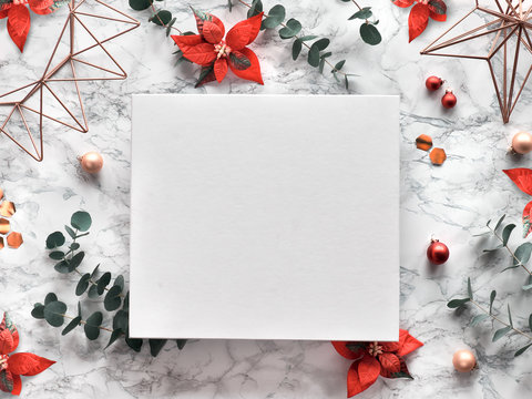 Christmas frame with fresh eucalyptus twigs, red poinsettia flowers, geometric decorations - hexagons, metal wire shapes. Trendy top view on white marble background. Copy-space on white canvas.