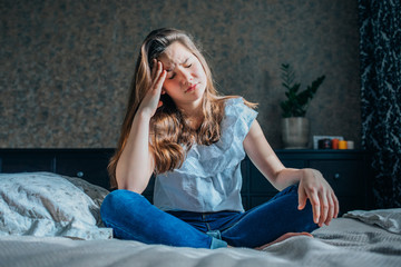 Obraz premium a sad dark-haired girl in a white blouse and blue jeans is sitting on the bed in her bedroom. She has a headache. horizontal shot, selective focus.