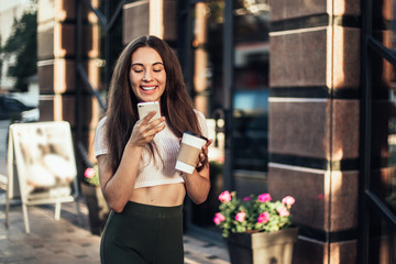 Smiling woman using phone and drink coffee on the street in summer day.