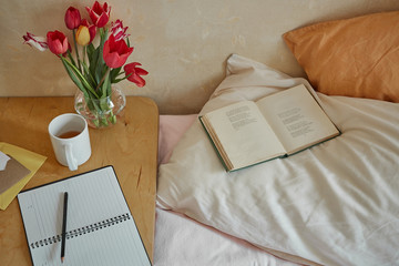 Wooden table with paper notebook, book, cup of tea and spring flowers next to bed