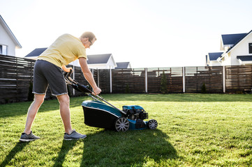 A young man is going to mow the lawn, he starts a push lawn mower. A guy in casual yellow t-shirt and in sunglasses - obrazy, fototapety, plakaty