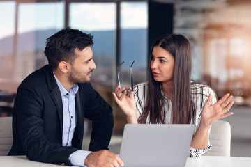 Couple of colleagues discussing work while sitting at the office and working on laptop