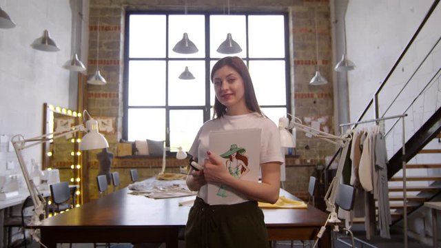 Zoom in of beautiful female designer standing in sewing workshop, holding album with fashion illustrations, looking at camera and smiling