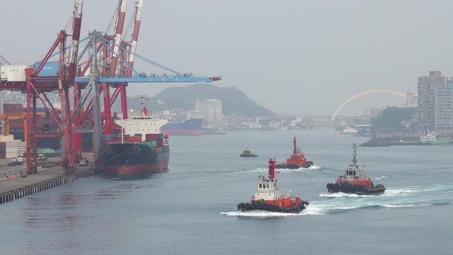 Three powerful orange tug boats sailing along waves of the ocean in  port with containers, cargo cranes, moored barges and barges against the backdrop of mountains and a big city on a foggy day