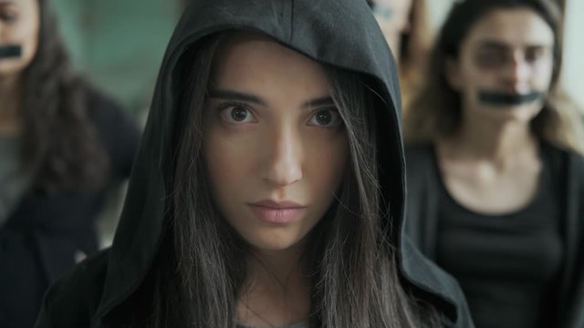 Unhappy girl in a black hood ,Sadly and depressively looking at the camera.They stand behind her,A young woman with bruises and blood on her face,