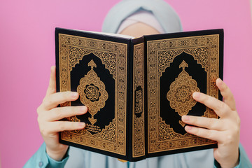 Muslim woman in a headscarf holds a Quran in her hands on a pink background
