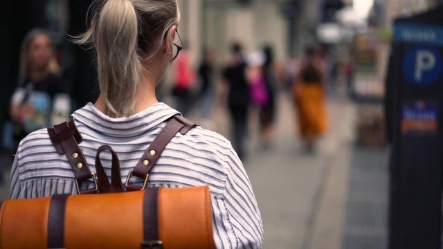 Back view of millennial hipster girl walking in slow motion on city street and taking pictures on modern smartphone camera. Female wanderlust with leather backpack exploring destination during trip 
