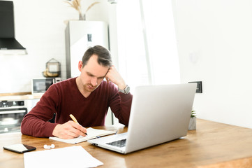 A guy uses laptop for distance study, he sits at the table at home and is writing lecture in a notebook