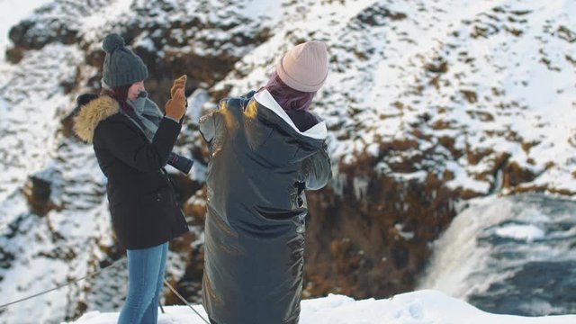 Two young beautiful girlfriends takes a photos on the background of a waterfall Skogafoss, Iceland