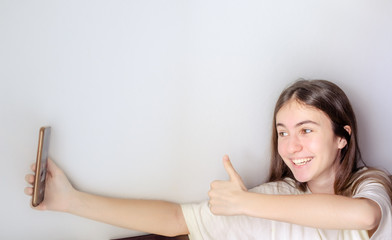 Smiling teen girl showing thumbs up symbol to the phone in hand on white gray background. Best friend chatting, video calling, social media, staying at home, quarantine, technology, modern lifestyle.