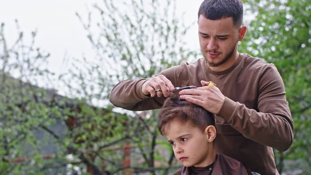 In the garden outside barber man make a haircut to the cute and sad small boy concentrated he cut hair with a scissors