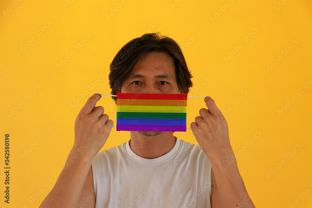 Wall mural a man with lgbt+ flag on hygienic mask in his hand and lifted up the front face on yellow background