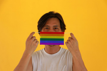 A man with LGBT+ flag on hygienic mask in his hand and lifted up the front face on yellow background. Tiny Particle or virus corona or Covid 19 protection.