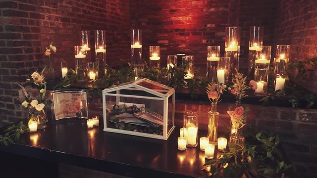 Transparent box for presents for groom and bride, wedding traditional decor elements on the table with lots of burning candles and floral decorative elements, dim roses and ruscus in front of brick wa