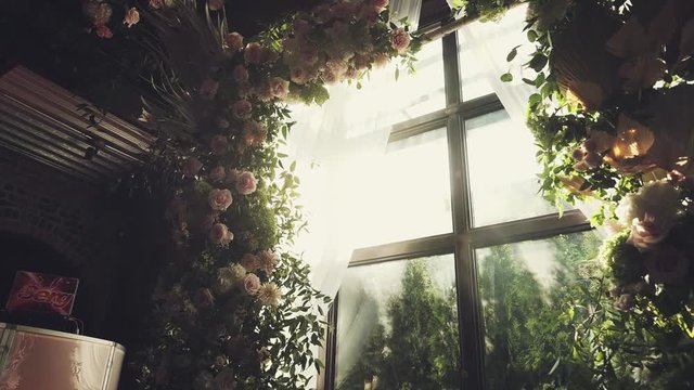 Luxury wedding ceremonial arch indoor near the huge window made of greenery and dim pink flowers, roses, peonies, ruscus
