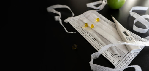with white medical objects located in a corner on a dark background, a protective medical mask with ties, pills, a thermometer, a glass of water, an apple. Copy space. Health care concept. 
