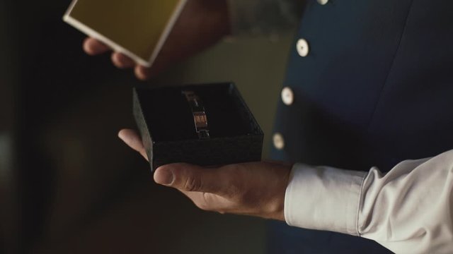 Front view of a man who is opening a small box with a wrist accessory, groom accessory, formal attire