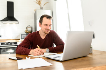 Male student writes a lecture in a notebook at home. He uses a laptop to watch a lecture.