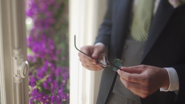 Man is holding eyeglasses in hands dressed in formal attire near the door, with view of violet flowers bush, front part view