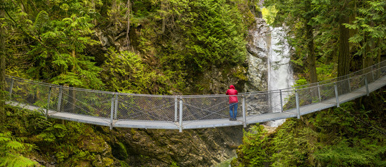 Panorama view of man in red hoodie standing on a suspension bridge and looking at the Cascade falls, in Cascade falls regional park, Deroche, British Columbia, Canada © Ferenc