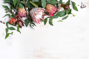 Beautiful pink king protea surrounded by  pink ice proteas, leucadendrons, eucalyptus leaves and flowering gum nuts, creating a floral border on a rustic white background. - obrazy, fototapety, plakaty