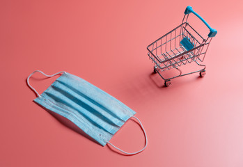 Medical mask and shopping trolley on coral background. The concept of lack of medical protection against infections and viruses. 