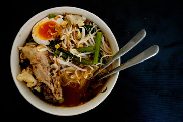 Noodle left view with egg ,vegetable ,Pork skeleton beef,spoon ,fork in green bowl and  black wood background with clipping path.Spicy noodle. Still life.nodle thailand food.