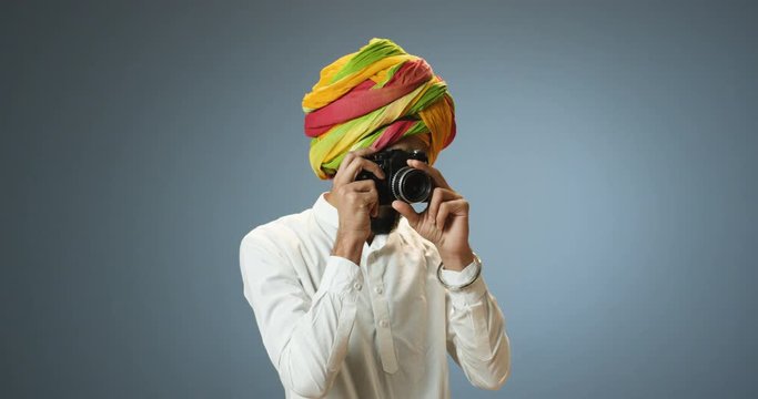 Young Hindu handsome man in traditional turban taking photos with vintage photocamera on gray wall background. Indian male making photographes with camera.