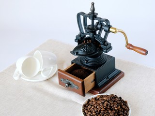 Coffee grinder, snow-white coffee cups and saucers, coffee beans close-up on the table surface.