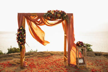 Square wooden wedding arch on outdoor sunset wedding ceremony. Red rose flowers and hanging cloth. Amazing warm sun light.