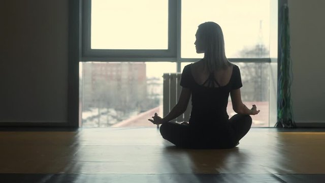 a woman meditates in the Lotus position in the training room in front of a large window. Slender athletic girl sitting on the floor. Exercise for concentration. the concept of a healthy lifestyle. Rel