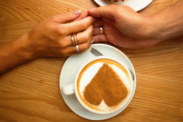 romantic lovely young couple in love sit in cafe having breakfast,closeup hands holding a cup of coffee with art design heart on it   on a wooden table background 