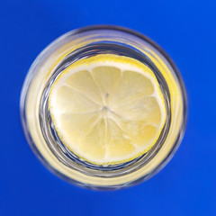 A slice of lemon in a transparent glass. Refreshing drink. Blue background. Top view. Square format.