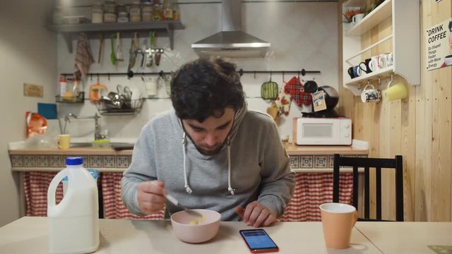 A young man having breakfast in the kitchen. Man eats Corn Flakes Cereal and uses smartphone