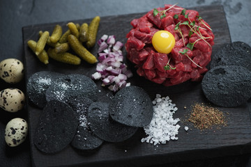 Black wooden serving board with beefsteak tartar and black potato chips, selective focus, studio shot