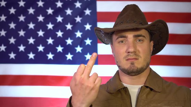 Independence Day, July 4th. A man in a cowboy hat and a leather jacket against the background of the American flag shows an indecent gesture, a phallic symbol.