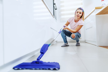 Attractive worthy caucasian blond young housewife crouching and cleaning kitchen floor with mop.