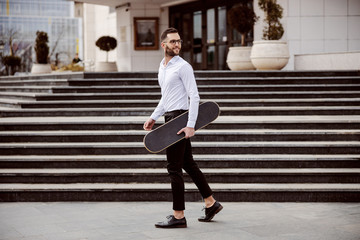 Full length of man in shirt walking outdoors and holding skateboard under armpit.