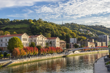 Fototapeta na wymiar Embankment by the Nervion River in Bilbao. Bend of the river. Colors of autumn in the city. Traveling in Spain. European city. Colorful cityscape by the river. Basque Country. The large city of Spain.