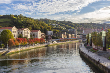 Naklejka na ściany i meble Embankment by the Nervion River in Bilbao. Bend of the river. Colors of autumn in the city. Traveling in Spain. European city. Colorful cityscape by the river. Basque Country. The large city of Spain.