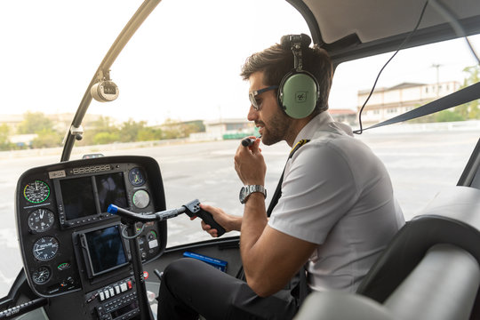 Picture of a male helicopter pilot in his white shirt with tie uniform. He is prepared for his flight. He sits inside a white helicopter with his head phone one ready to take off.